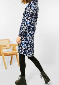 Kobieta w niebieskiej, białej i czarnej sukience w zwierzęcy wzór, czarnych legginsach i czarnych butach z sznurówkami, przechodzi obok drewnianego krzesła.