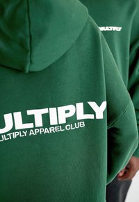 Sudadera de color verde oscuro hecha de una mezcla de algodón. Presenta un gran texto en blanco con el logo "MULTIPLY" y "MULTIPLY APPAREL CLUB" en la parte posterior.