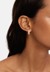 Złote kolczyki w kształcie obręczy z wieloma rzędami małych białych koralików, podkreślające elegancki design. Noszone na uchu modelki, ukazujące profil boczny.