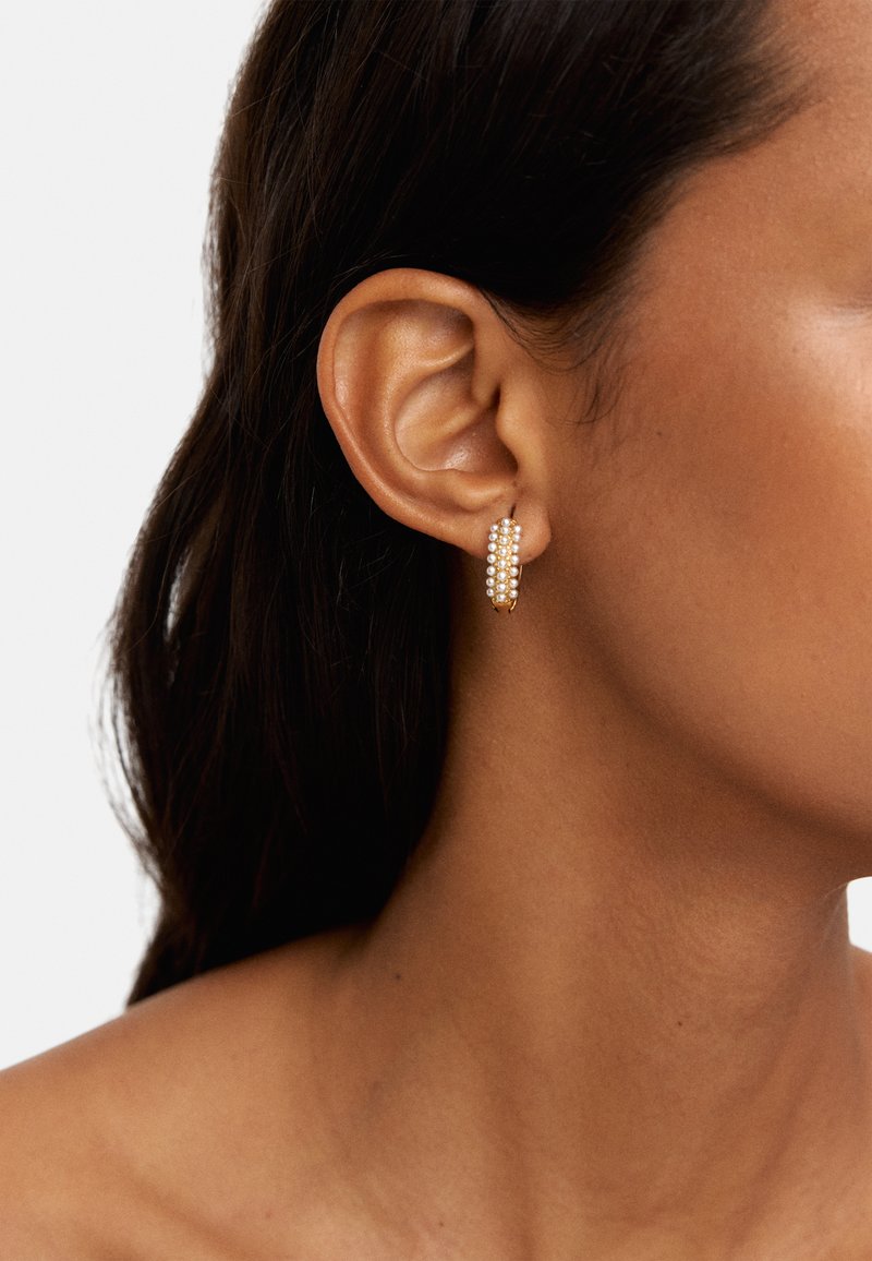 Złote kolczyki w kształcie obręczy z wieloma rzędami małych białych koralików, podkreślające elegancki design. Noszone na uchu modelki, ukazujące profil boczny.