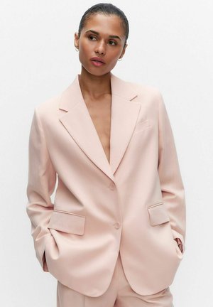 Vrouw met haar in een knot, gekleed in een lichtroze oversized blazer en bijpassende broek, staand met handen in de zakken tegen een effen achtergrond.