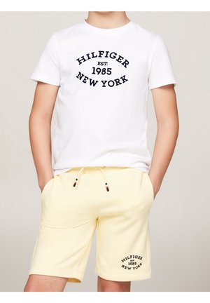 Dziecko noszące białą koszulkę i jasnżółte szorty z tekstem "Hilfiger Est. 1985 New York" nadrukowanym na obu ubraniach.