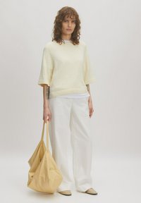 Cremiger Pullover mit Struktur, darunter ein weißes, geschichtetes Hemd, weite weiße Hosen und eine gelbe, lässige Tasche. Das Modell steht vor einem neutralen Hintergrund.