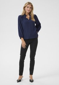 Blusa in blu navy con texture, scollatura a V e dettagli arricciati sulle maniche, abbinata a pantaloni neri slim-fit e mules nere con tacco.