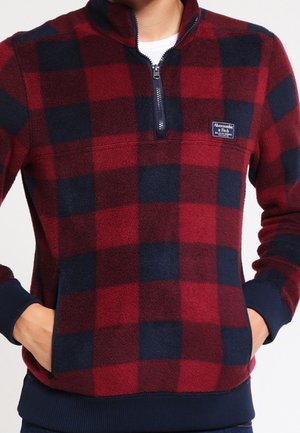 Person wearing red and navy blue checkered fleece pullover with half zipper and hands in pockets.
