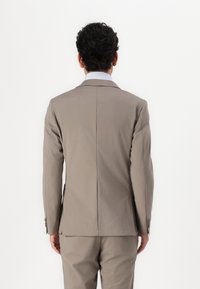 Jachetă de costum maro deschis cu o textură netedă, design structurat, rever decupat și fante unice la spate. Croială ajustată și detalii minime.