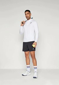 Camisa de polo blanca de manga larga, pantalones cortos negros, zapatillas blancas y calcetines a rayas. Sosteniendo una raqueta de tenis y una pelota. Atuendo atlético casual.