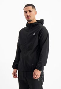 Zwarte zip-up jack met capuchon, met een slank ontwerp, elastische manchetten en subtiele logo-accenten. Het materiaal lijkt lichtgewicht en glad.