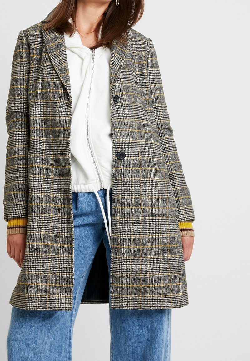 Femme portant un manteau à carreaux gris par-dessus un sweat à capuche blanc zippé et un jean bleu, les mains détendues le long du corps sur un fond uni.