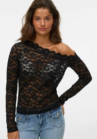 Zwarte kanten top met lange mouwen, een asymmetrische halslijn, ingewikkelde bloemenpatronen en een semi-doorzichtige textuur. Gecombineerd met blauwe jeans.