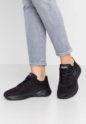 Person trägt hellgraue Jeans und schwarze Skechers Arch Fit-Sneaker, steht auf weißem Boden, dargestellt von den Waden abwärts.
