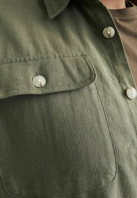 Cămașă din țesătură verde cu un buzunar cu nasturi, având nasturi mari și de culoare deschisă. Textura pare netedă, cu variații subtile de culoare.