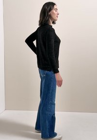 Femme en profil portant un pull noir à manches longues, un jean bleu et des baskets blanches, debout devant un mur uni de couleur claire.