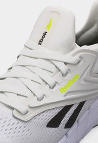 Άσπρο αθλητικό παπούτσι Reebok με άσπρα κορδόνια, νεονί κίτρινο λογότυπο Reebok στο πλάι και στη γλώσσα, μαύρη λεπτομέρεια στο πάνω μέρος από διχτυωτό υλικό και υφή στη σόλα.