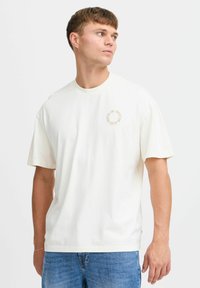 Light beige crew-neck t-shirt in soft cotton, featuring a circular printed text detail in light brown on the left chest area.