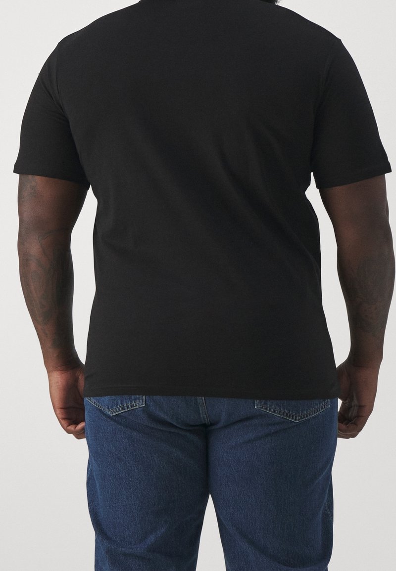 Camiseta negra de manga corta con escote redondo, mostrada desde la parte trasera, combinada con jeans de mezclilla azul. La tela parece suave y transpirable.