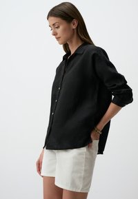 Camicia nera in lino con colletto, vestibilità leggermente rilassata e maniche arrotolate. Abbinata a pantaloni corti di jeans bianchi.