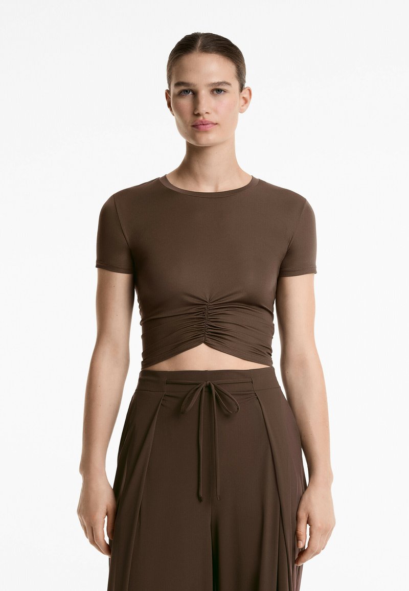 Maglietta cropped marrone realizzata in tessuto liscio, con scollo rotondo e maniche corte. Il dettaglio centrale arricciato crea una forma aderente.