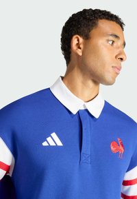 Polo blu con colletto bianco, presenta un logo di gallo rosso e tre strisce bianche sulle maniche. Realizzata in tessuto liscio, con una vestibilità classica.