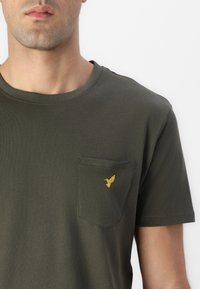 Homme portant un t-shirt vert foncé avec un petit emblème jaune d'oiseau sur la poche gauche de la poitrine, sur un fond blanc uni.