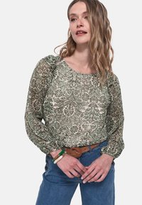 Blusa verde a fantasia floreale con scollatura rotonda e maniche a sbuffo, abbinata a jeans blu e una cintura marrone. Tessuto leggero e texturizzato.