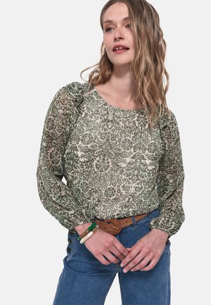 Blouse verte à motif floral avec un col rond et des manches bouffantes, associée à un jean bleu et une ceinture marron. Tissu léger et texturé.