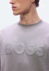 Γκρι φούτερ με ανάγλυφο, υφασμάτινο λογότυπο "BOSS". Διαθέτει ριμπ λαιμόκοψη και λείο υφασμάτινο φινίρισμα. Κοντινή λήψη.