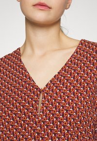 Gros plan d'une personne portant un chemisier rouge à motifs circulaires et géométriques, avec une petite ouverture nouée devant.