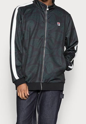 Zwarte jas met ritssluiting en donker paisley-patroon, witte strepen op de mouwen en een klein Fila-logo op de linkerkant van de borst, gedragen met donkere spijkerbroek.