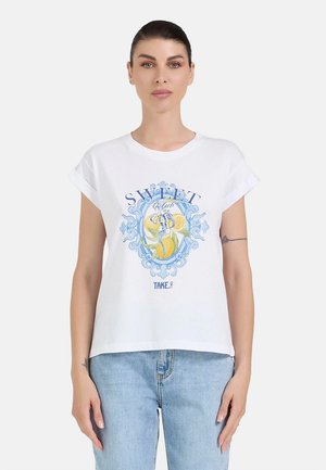 Donna con capelli scuri con una T-shirt bianca con maniche arrotolate con un motivo di limoni blu e gialli e jeans azzurri chiari, in piedi davanti a uno sfondo semplice.
