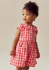 Bambino piccolo che indossa un vestito rosso e bianco a quadri con bottoni e tasche, che guarda di lato con capelli ricci.