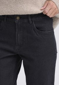 Juodos spalvos džinsai iš džinso audinio, klasikinio kirpimo, turintys priekines kišenes, žalvarinį sagos užsegimą ir subtilias siuvimo detales. Audinio paviršius tekstūruotas.