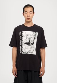 Tânăr purtând un tricou mare, negru, cu un grafic alb al unei păsări și textul "Versace Jeans Couture" pe față, stând în fața unui fundal simplu.