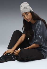 Beanie grigio chiaro con design di animali neri, pantaloni neri larghi e una maglietta grigio scuro con un motivo sottile e logo. Sneakers nere.