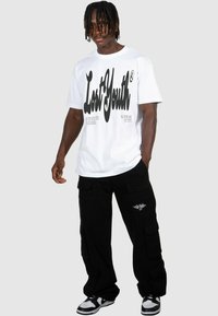 Vit grafisk t-shirt med fet svart text, i kombination med svarta cargo-byxor med flera fickor och en liten vit logotyp på benet.