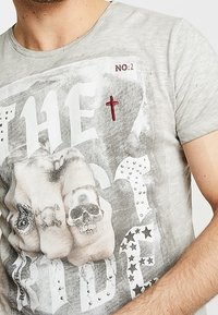 Camiseta de jersey gris con un estampado gráfico que muestra manos con tatuajes, calaveras y tachuelas decorativas; cuello redondo; textura suave.