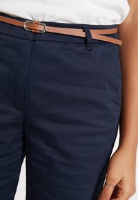 Hnědý kožený pásek se stříbrnou D-kroužkovou sponou, nošený přes námořnické kalhoty. Tkanina má hladkou strukturu s viditelnými záhyby.