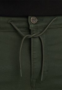 Groene broek van gestructureerde stof, met een trekkoordsluiting en een metalen knoopaccent. Close-upweergave die de details en kleur benadrukt.
