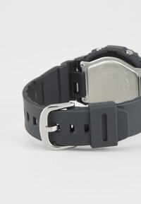 Bracelet de montre en silicone noir avec une boucle en métal argenté, comportant des trous découpés pour ajustement, et un dos métallique avec des détails en relief.
