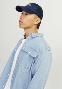 Gorra de béisbol navy con parte trasera de malla, llevada con una camisa de denim azul claro que tiene un bolsillo en el pecho y cuello abotonado sobre una camiseta blanca.