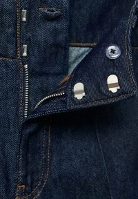 Details van een spijkerjack met een gedeeltelijk open rits aan de voorkant, koperen stiksels, zilveren hardware en een donkerblauwe stoftextuur.