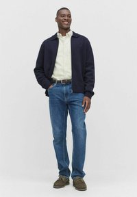 Leende man iklädd en mörk marinblå jacka, krämfärgad skjorta, blåa jeans, brunt bälte och bruna skor, stående med ena handen i fickan.