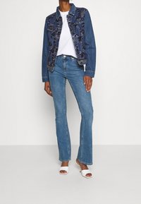 Jeansowa kurtka z haftowanymi detalami, zestawiona z jasnoniebieskimi spodniami dzwonami i białymi szpilkami na odsłoniętych palcach. Pod spodem prosta biała koszulka.