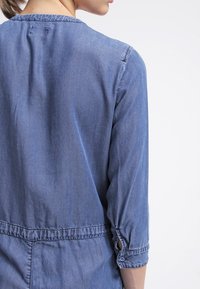 Jeansklänning i ljusblått, med trekvartsärmar och knappar på ärmsluten. Har en insvängd midja och en mjuk, sl ät textur.