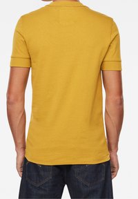 Mosterdgele T-shirt met korte mouwen, ronde hals en een effen textuur, met een klein vierkantembleem op de achterkant.