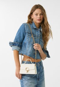 White mini handbag with a gold ring hardware detail, worn with a cropped denim jacket and high-waisted blue jeans.