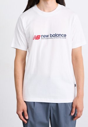 Osoba w białej koszulce New Balance z czerwonym i czarnym logo oraz napisem, w połączeniu z niebieskimi spodniami.
