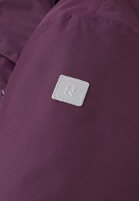 Tessuto viola con una consistenza liscia, caratterizzato da una toppa quadrata grigia con il logo 