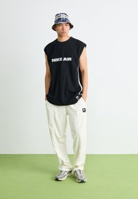 Moški v brezrokavniku črne barve Nike Air, belih hlačah Nike Air, karirasti klobuk iz blaga in supergah stoji na zeleni podlagi ob belem zidu.