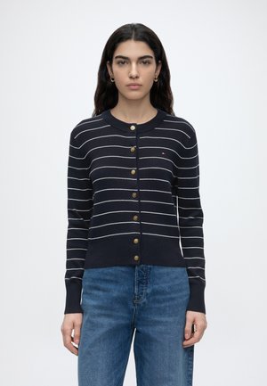 Femeie cu păr lung și închis la culoare poartă un cardigan bleumarin cu dungi albe și nasturi aurii, asortat cu blugi albaștri din denim, stând pe un fundal alb.